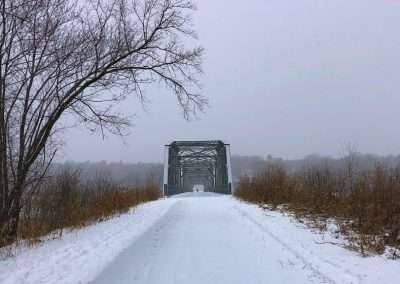 A metal truss bridge sits at the end of a snowy footpath