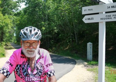A man with a white beard and a helmet poses near a sign along a trail