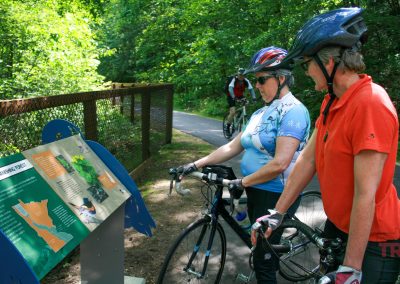 Two women wearing helmets study an interpretive sign along a trail