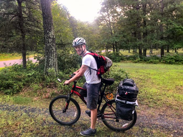 Sam Marsnik A man wearing a helmet and backpack straddle his bicycle, which is packed with panniers.