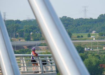 A woman wearing a helmet pauses to take in the view of a river from a tall bridge