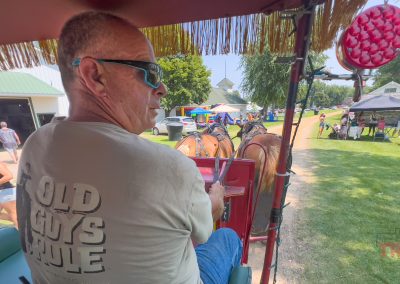 A man wearing sunglasses drives a team of horses pulling a wagon at a fair