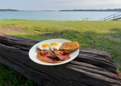 A plate of breakfast food sits on a log by a lake
