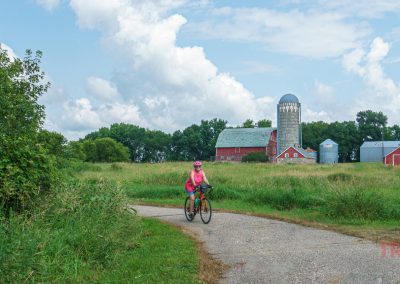 A woman rides her bike on a paved trail past a red barn and concrete silo
