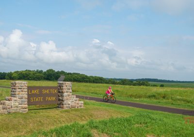 A woman rides her bike on a paved trail past the brown sign for Lake Shetek State Park
