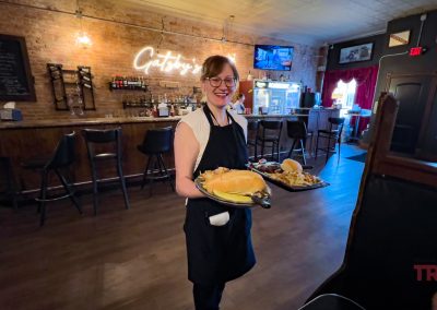 Smiling server in a black apron presents a tray with a large sandwich and fries inside the cozy bar adorned with wooden decor. A neon sign reading Gatsbys glows on the brick wall, evoking the charm of a rustic retreat after exhilarating bike rides through winding trails.