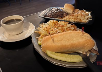 A plate with a sandwich, pickles, and hash browns is in the foreground, while another with steak and hash browns sits beside it, offering the perfect meal after exploring Minnesota trails. A cup of black coffee is placed on the left side of the table.