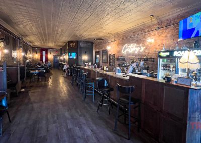 A cozy bar interior with a long wooden counter, high chairs, and exposed brick walls. A neon sign reads Craftys above the bar, welcoming patrons after a day on the bike trails. Seating lines the wall as intricate tin detailing adorns the ceiling and a TV is mounted nearby.
