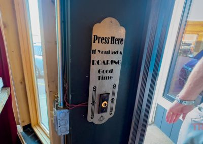 A vertical sign by the door reads Press here if you had a roaring good time, with a button below. A persons arm is visible near the sign, as though they just returned from exhilarating Minnesota trails, and part of a car can be seen outside through the window.