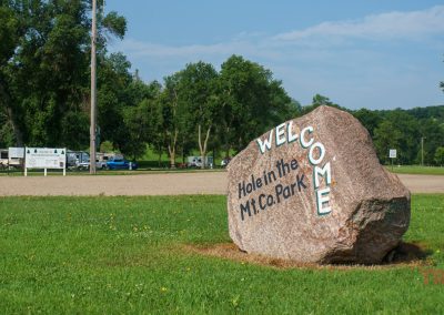 A large stone with the words Welcome Hole in the Mt. Co. Park stands on grass, inviting visitors to explore the nearby bike trails. In the background, parked RVs and trees bask under a clear blue sky.