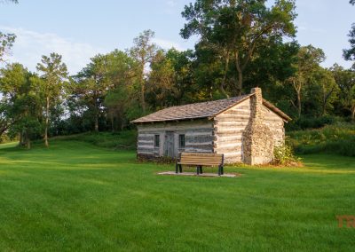A small, rustic log cabin stands in a lush green field surrounded by trees, perfect for starting your Minnesota bike rides. A wooden bench sits out front as the sky stretches clear with a few clouds.