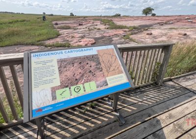 An interpretive sign sits on a wooden platform at the edge of a red rock plateau