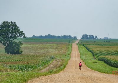 A woman rides her bike on a gravel road flanked by corn and soybean fields