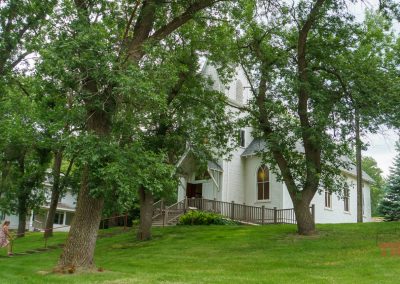 A woman walks toward a white church on a lwan with trees