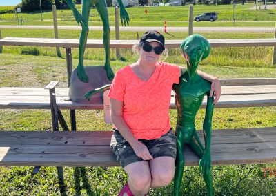 A woman sits on a bench with her arm around a statue of a green alien figure