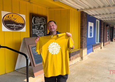 A man holds up a yellow t-shirt featuring a sunflower and the words 'mountain lake'.