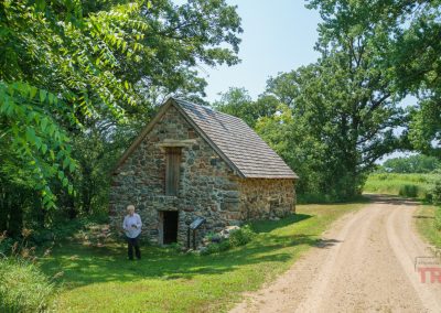 A small house made of field stone sits in a clearing of a forest by a gravel road
