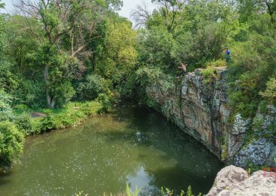 A boy jumps into a pond from a cliff while a man records it on his cell phone