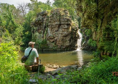A man with a backpack sits on a rock near a pond with a waterfall