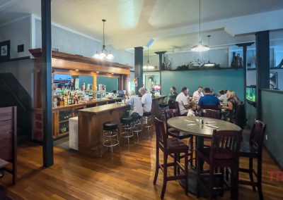 Patrons sit at a table and a bar in a restaurant with a white wood ceiling