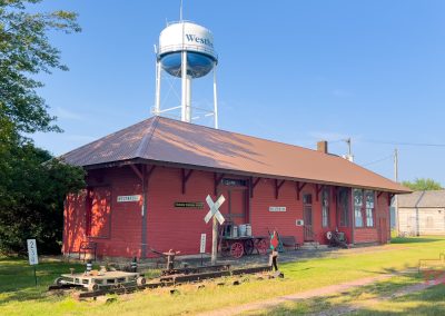 Exterior view of a historic train depot with red wood siding, track remnants and a water tower in the background