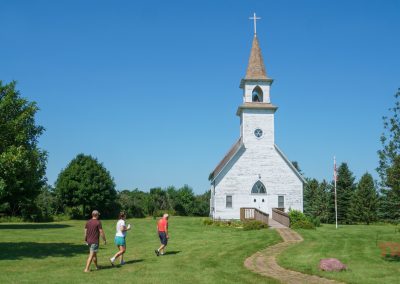 Three people walk toward a historic wood clad church on a grassy field