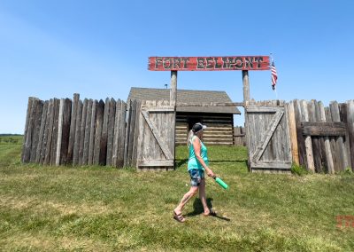 A woman wearing shorts walks by a stockade signed "Fort Belmont"