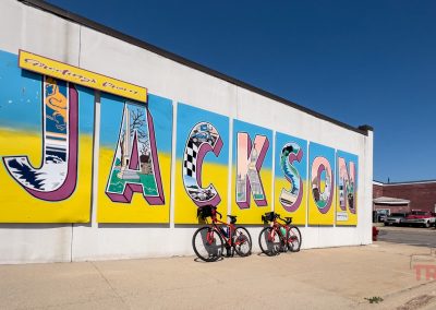 Two bicycles lean against a wall with a mural that reads "Greetings from Jackson"