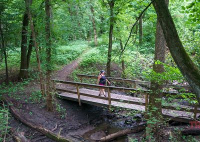 A woman walks across a bridge in a forest at Kilen Woods State Park.