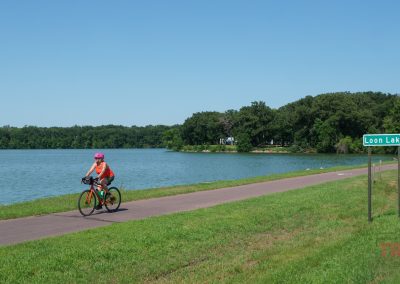 A woman rides her bike on a paved trail past a lake