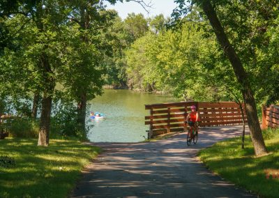 A woman rides her bike on a paved trail past a lake