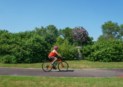 A woman rides her bike on a paved trail past an art display made of bike wheels