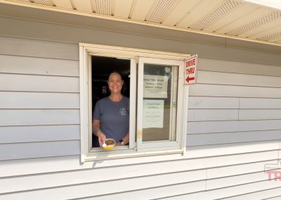 A woman poses for as picture at a drive through window holding a small tin of cheesecake