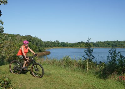 A woman rides a mountain bike through a grassy field past a lake at Kensington Rune Stone Park.