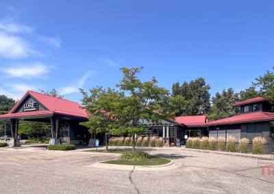 A building with a red roof and a sign reading Lore nestles among the Minnesota greenery. The foreground boasts an empty paved parking area under a blue sky, perfect for starting bike rides. The word trails is faintly visible on the lower right.