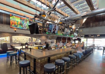 A spacious bar area with high ceilings and exposed beams, reminiscent of a mountain lodge. The central counter is surrounded by blue cushioned stools, TV screens, and colorful decor. Large windows let in natural light, like the serene aura of nearby hiking trails. Two women sit at the bar as a bartender serves them.