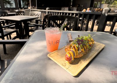 A loaded foot-long hot dog topped with colorful sauces and greens on a wooden board accompanies a pink cocktail with a straw. Set on a gray table in Minnesota, the outdoor patio offers views reminiscent of nearby bike trails with empty black chairs and a fence in the background.