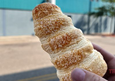 Close-up of a hand holding a flaky, sugar-dusted pastry against an urban background. The slightly blurred backdrop, reminiscent of serene Minnesota landscapes, features a teal building and a tree.