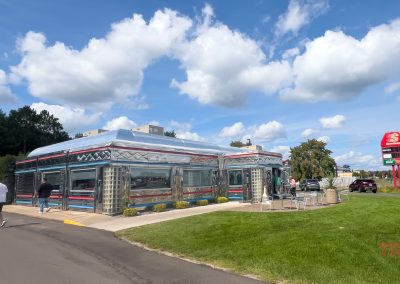 A retro-style diner with a chrome exterior, large windows, and red and blue trim sits near the popular Paul Bunyan Trail. People stroll nearby, while cars are parked in the background by a red and black sign. The sky is partly cloudy, and a well-maintained lawn surrounds the diner.