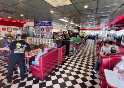 A busy diner with black and white checkered floors, red booths, and vibrant decor. Perfect for refueling after exhilarating bike rides. People are seated and a server in black delivers food. Walls are adorned with signs and memorabilia. The atmosphere is lively and bustling.