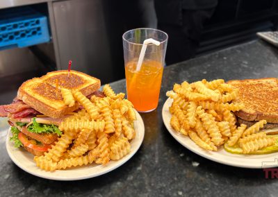 Two plates of crinkle-cut fries with sandwiches sit on a dark countertop. One sandwich has layers of bacon, lettuce, and tomato, perfect fuel after a day exploring bike trails. A glass of orange drink with a straw is in the background. A pickle slice accompanies the plate on the right.