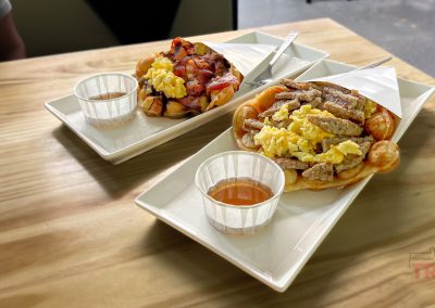 Two plates with waffles topped with scrambled eggs, sausage, and bacon sit on a wooden table. Each has a small cup of syrup beside it. This hearty meal awaits after exploring Minnesotas scenic bike trails, blending excitement with savory satisfaction.