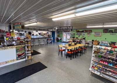 A spacious, brightly lit dining room with a vintage decor. There are booths and a long counter on the left, tables with chairs in the center, and racks of snacks on the right. A sign reading The Gathering Place welcomes those fresh from invigorating bike rides.