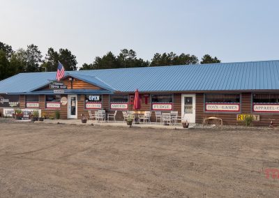 Single-story roadside store with signs for coffee, ice cream, sweets, and gifts. Blue metal roof, wooden exterior, and an American flag. Nestled among trees with a dirt parking lot out front, it's a perfect stop for those exploring the nearbyPaul Bunyan Trail on a slightly overcast day.