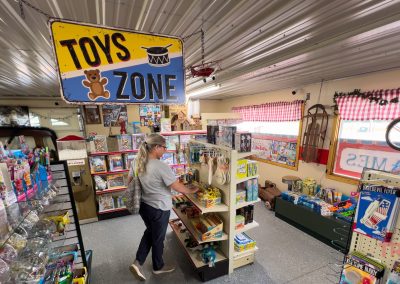 A person with long gray hair browses a shelf filled with toys in Toys Zone. The room, has a metal ceiling, vintage decor, and red and white checkered curtains covering the windows.