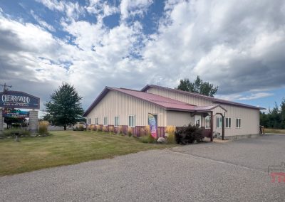A light beige building with a brown roof sits beside the road. A dark sign reads Cherrywood Hand Crafted Fabrics. The sky is partly cloudy, and trees surround the area. The ground is asphalt and grass.