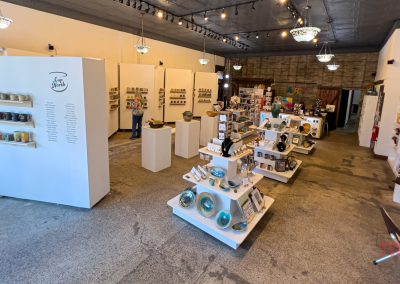 A pottery store interior with ceramic items displayed on white shelves and panels. Mugs, plates, and other pieces are neatly arranged. Decorative lighting fixtures hang from the ceiling, while artworks decorate the walls