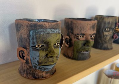 Four ceramic mugs with sculpted faces, each featuring unique colors and expressions, are lined up on a wooden shelf. The mugs have a textured surface and display faces with intricate details and vibrant hues.