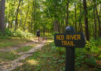 Trail sign at Crow Wing State Park reading Red River Trail is set in a lush, green forest. A person wearing a backpack is seen walking along the dirt path in the background.