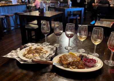 A restaurant table with two plates of food garnished with herbs and two sets of tasting glasses filled with different colored drinks. The background shows a dimly lit dining area with a few people sitting at tables.